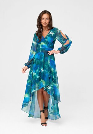 Vrouw draagt een blauwgroene bloemige high-low jurk met lange open mouwen, staand met één hand in haar taille en zwarte hakken.