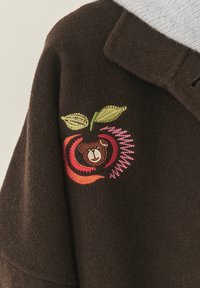 Barna gyapjú keverék kabát, amely egy színes hímzett mintát ábrázol: egy alma belsejében egy medve, zöld levelekkel és rózsaszín részletekkel.