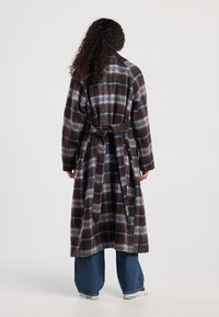 Long plaid coat in brown, blue, and white, made of a soft material, featuring a tie waist and relaxed fit. Visible oversized pockets.