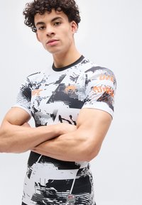 Νέος άνδρας με σγουρά μαλλιά στέκεται με σταυρωμένα τα χέρια, φοράει στενό μαύρο και λευκό μπλουζάκι προπόνησης UFC με πορτοκαλί λογότυπα και αφηρημένα σχέδια.