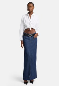 Denim maxi skjørt med en frontsplitt, befestet med et svart belte. Kombinert med en knyttet hvit skjorte, som skaper et uformelt lagdelt utseende.