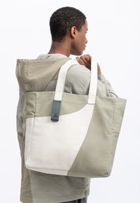 Tote bag met een gesplitst ontwerp in lichtgroen en wit, voorzien van stevige handvaten en een gestructureerde, geweven stof. Versterkt met een grijze label.