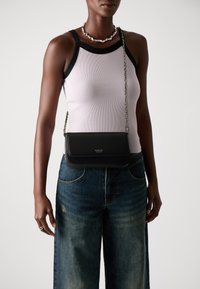 Vrouw in een mouwloze witte top met zwarte rand, blauwe denim jeans, een zilveren kettinghalsketting en een zwarte schoudertas met kettingriem.