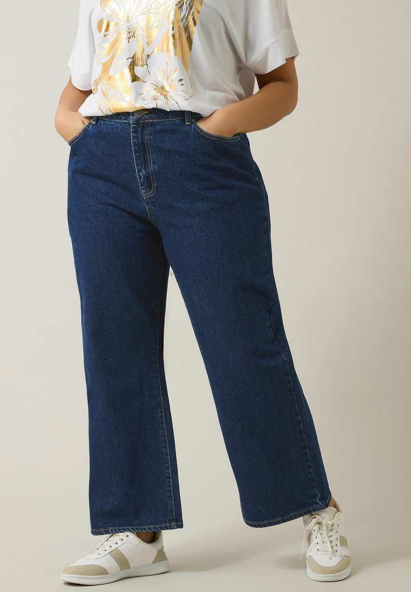 Jeans de mezclilla azul oscuro de pierna ancha con bolsillos cosidos y cintura alta, combinados con zapatillas blancas con acentos beige.