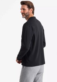 Schwarzes Langarm-Poloshirt mit geradem Kragen, getragen mit hellgrauen Hosen. Glatte Textur mit taillierter Passform. Keine sichtbaren Muster.