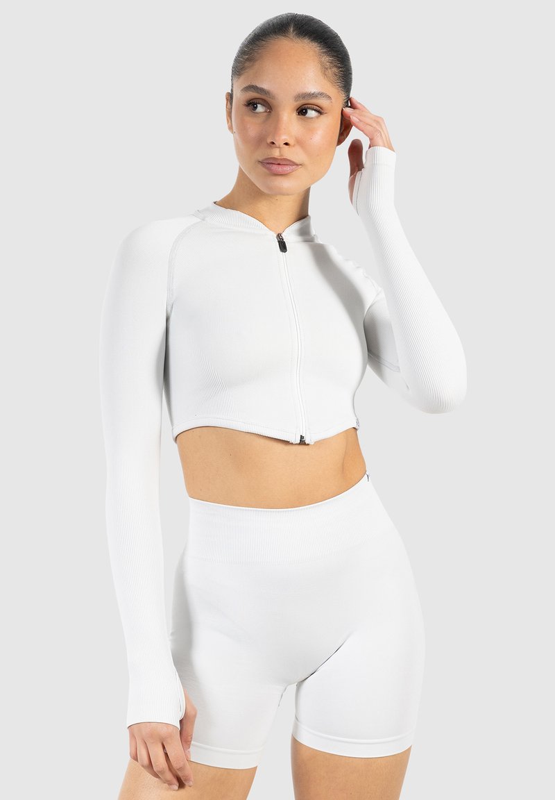 Hvid cropped zip-up langærmet top med ribbet tekstur, parret med højtaljede hvide shorts. Simpelt design, elastisk stof.