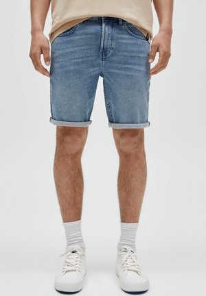 Homme portant un short en denim bleu retroussé, des chaussettes blanches et des baskets blanches, debout devant un fond clair uni.