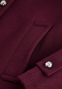 Close-up van een bordeaux wollen jas met een zakflap en mouwmanchet, versierd met een zilverkleurige decoratieve knop.
