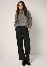 Szary sweter z golfem o fakturowanej strukturze w połączeniu z luźnymi czarnymi spodniami z elastycznym pasem. Model stoi z rękami w kieszeniach.