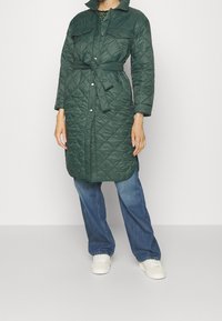 Mujer vestida con un abrigo acolchado verde de longitud media y cinturón, sobre jeans azules y zapatillas blancas, de pie contra un fondo claro y liso.