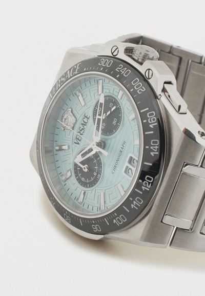 Ασημένιο ρολόι χειρός chronograph Versace με ανοιχτό μπλε καντράν, μαύρους υποκαντράν, στεφάνη ταχύμετρου, παράθυρο ημερομηνίας και μπρασελέ από ανοξείδωτο ατσάλι.