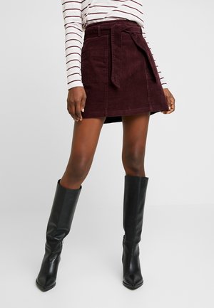 Femme portant une mini-jupe en velours côtelé bordeaux avec une ceinture nouée, un haut à manches longues rayé et des bottes en cuir noires montantes jusqu'aux genoux, debout sur un fond clair.