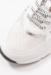 Bílé tenisky s perforovanou kůží na špičce, stříbrnými akcenty a černo-bílými šňůrkami, zobrazené na bílém pozadí.