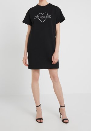 Vrouw die een korte zwarte jurk draagt met de tekst "LOVE MOSCHINO" binnen een hart van strass-steentjes, gecombineerd met zwarte sandalen met hoge hakken.