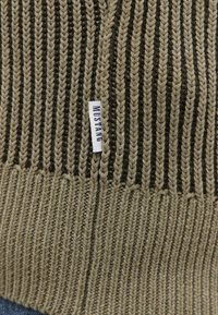 Adītā beanie olīvzaļā un tumši brūnā krāsā ar vertikālu ribu tekstūru. Tam ir mazs balts birku ar uzrakstu "MUSTANG". 