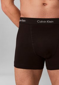 Männlicher Torso mit schwarzen Calvin Klein Boxershorts und Logo auf dem Bund vor grauem Hintergrund.