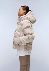 Beige vadderad pufferjacka med huva, med ett rutmönster och dragkedja fram. Dekorerad med ett logopatch på ärmen.