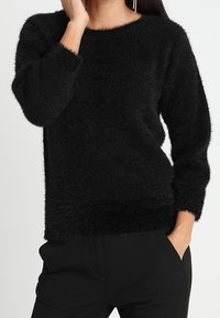 Pull noir en pilou avec un col rond et des manches longues. La texture semble douce et duveteuse, offrant une coupe décontractée au niveau des hanches.