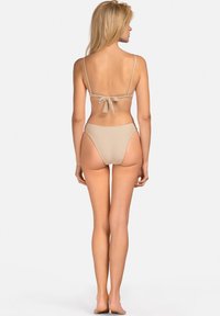 Kobieta z blond włosami stoi boso, zwrócona plecami, w beżowym bikini z zawiązywanym na plecach topem i wysokim stanem majtek na gładkim tle.