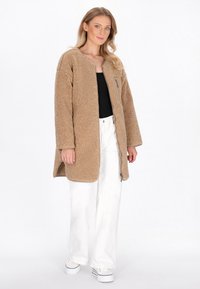Donna con cappotto in pile beige con tasca con zip sopra una maglietta nera, pantaloni bianchi a gamba larga e sneakers platform bianche, in piedi su uno sfondo bianco.