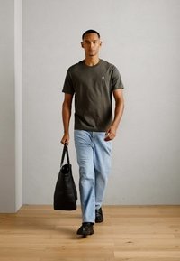 Black leather tote bag, lightweight olive green tee, light blue denim jeans, black shoes; model walking on wooden floor against a simple wall.