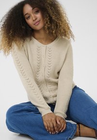 Beige knitted cardigan with a V-neck, featuring button closure and delicate lace patterns along the sleeves and front. Casual fit, paired with blue denim.