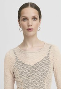 Blusa in pizzo di un leggero color beige con un motivo floreale, dal design aderente e scollatura a barchetta. Le maniche lunghe e le parti trasparenti mettono in risalto la texture.