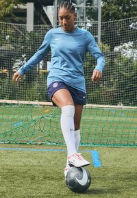 Kék, hosszú ujjú sportpóló fehér díszítéssel, tengerészkék sorttal és fehér térdig érő zoknival. Egy focilabda van a lábán mesterséges fűben.