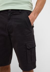 Mężczyzna w czarnych szortach cargo z boczną kieszenią i ręką w przedniej kieszeni, w zestawie z jasnozieloną koszulą.