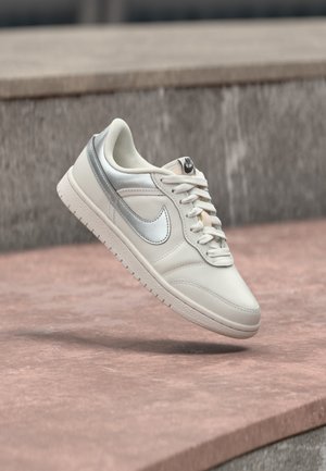 Biela teniska so strieborným logom Nike a šnúrkami, zobrazená vo vzduchu nad kamenným povrchom a rozmazaným kamenným pozadím.