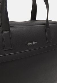 Schwarze Lederhandtasche mit glatter Textur, strukturierter Form und eing geprägtem Logo auf der Vorderseite. Zwei stabile Tragegriffe.