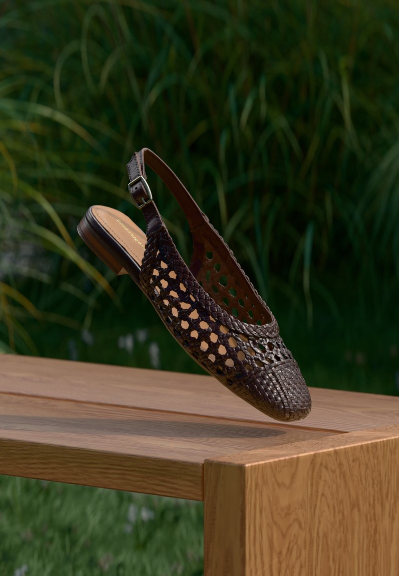 Mørkebrunt flettet læder slingback-fladsko svæver over en træoverflade med grønne planter sløret i baggrunden.