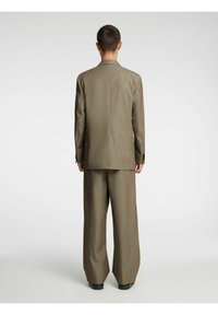 Brūns pielāgots uzvalks ar strukturētu jaku un platām biksēm. Audums izskatās gluds ar smalku rakstu un pogu akcentiem.