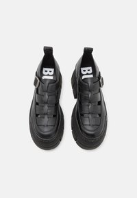 Crne slip-on cipele od teksturirane kože, sa tkanim dezenom, debelim gumastim đonom i podesivim detaljem sa kopčom. Bijeli logo vidljiv.