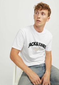 Camiseta blanca de algodón con un logo gráfico negro "JACK&JONES" con acentos dorados. Diseño de manga corta y cuello redondo.