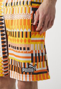 Moniväriset urheilushortsit, joissa on rohkeita geometrisiä kuvioita oranssissa, keltaisessa, mustassa ja valkoisessa; mukana Puma-logo helmassa.