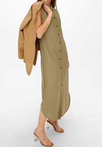 Donna in un vestito midi senza maniche di colore verde oliva, che tiene una giacca marrone chiaro sulla spalla, indossando sandali con tacchi trasparenti.