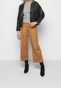 Black leather jacket with zippered pockets, gray turtleneck, and tan wide-leg trousers with rolled cuffs, paired with black ankle boots.