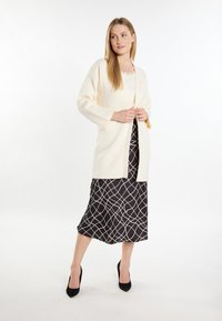 Cremefarbener Cardigan mit langen Ärmeln und offenem Vorderteil, getragen über einem schwarzen Kleid mit einem weißen geometrischen Muster. Schwarze Absätze vervollständigen das Outfit.