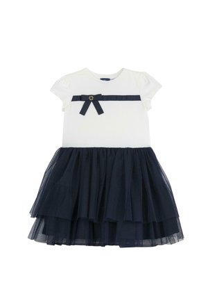 Biały bawełniany top z krótkimi rękawami, granatowa spódnica z warstwami tiulu, granatowa wstążka z kokardą w talii oraz okrągły złoty akcent.