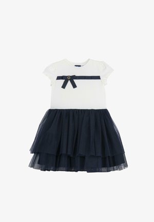 Hvid bomuldsbluse med korte ærmer, navy tyl lagdelt nederdel, navy bånddetalje med sløjfe ved taljen og rund guldfarvet accent.