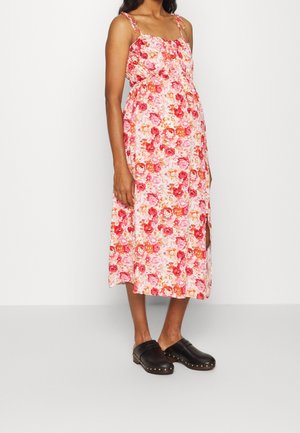 Frau trägt ein rosa Midikleid mit Blumenmuster und Spaghettiträgern sowie schwarze, mit Nieten verzierte Slipper, steht vor einer schlichten weißen Wand.