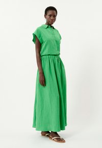 Green linen dress with short sleeves, button-up front, elastic waist, and long flowy skirt. Paired with flat sandals. Simple design.