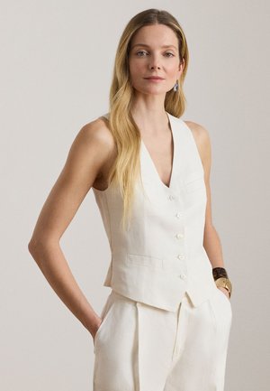 Kvinde med langt blondt hår iført en hvid ærmeløs knapvest og matchende bukser, hænderne i lommerne, mod en ensfarvet lys baggrund.