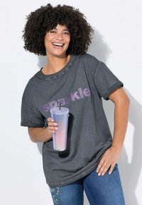 Grå oversized t-shirt med "sparkle" i lilla glimmer, opsmøgede ærmer, parret med denim jeans og holder en holografisk tumbler.