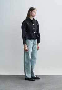 Vrouw staand in zijaanzicht, gekleed in een zwart jasje met zilveren knopen, lichtblauwe jeans met wijde pijpen en zwarte loafers op een grijze vloer.