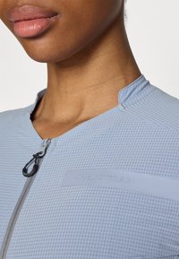 Hellblauer Zip-Up-Top aus strukturiertem Stoff mit Karomuster, Stehkragen und rechteckigem Logo-Patch auf der Brust.