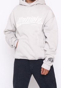 Felpa con cappuccio grigio chiaro, dotata di tasca frontale, un audace logo "Multiply", polsini a costine e una vestibilità rilassata. Abbinata a pantaloni scuri.