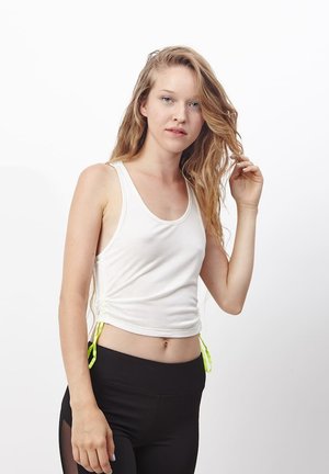 Joven mujer con cabello largo y ondulado, vistiendo una blusa sin mangas blanca y leggings negros, tocándose el cabello frente a un fondo blanco liso.