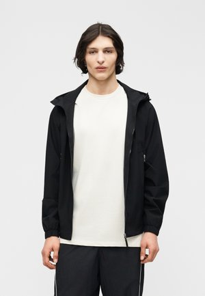 Jonge man met halflang donker haar, gekleed in een wit T-shirt, een zwart jack met ritssluiting en capuchon, en zwarte broek met witte strepen.